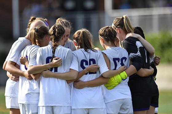 Photo of Chatham University's women's soccer team in white and purple uniforms huddles on the field during a match. 