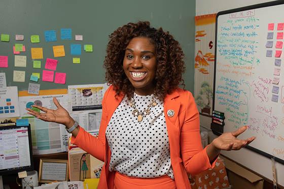 Photo of a Chatham University MSUS+MBA graduate in a professional suit, posing in front of her desk with a white board and post-it notes in the background