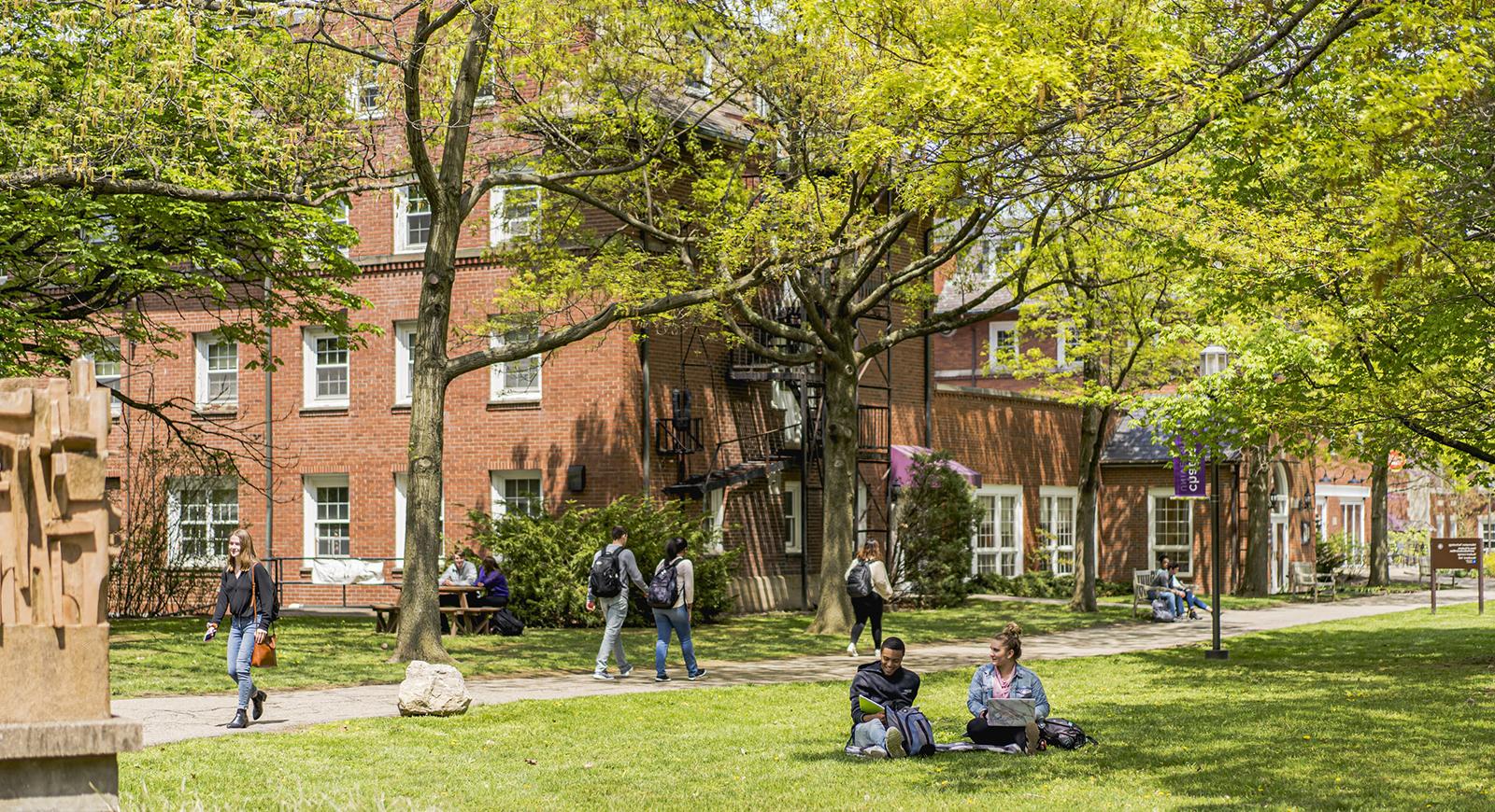 Photo of Chatham University students walking to class and sitting outside on the academic quad on a sunny day surrounded by red-brick academic buildings and greenery