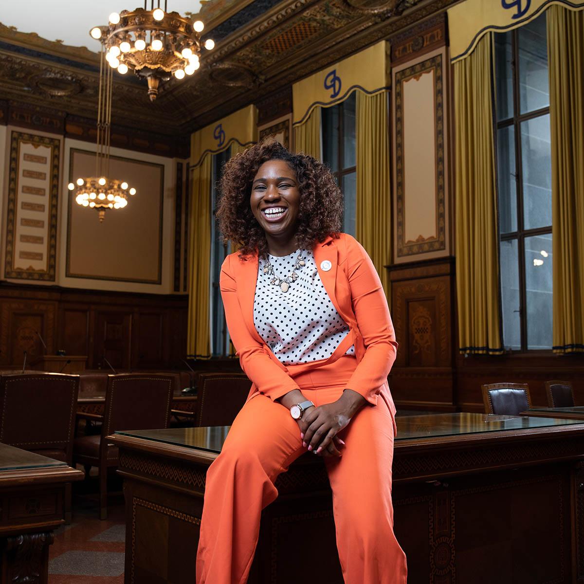 Photo of Feyisola, a young Black woman in a bright suit, poses smiling and laughing against a table in an ornate room