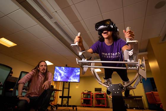 Photo of a student wearing a purple Chatham University shirt, in a virtual reality machine, while a professor watches on